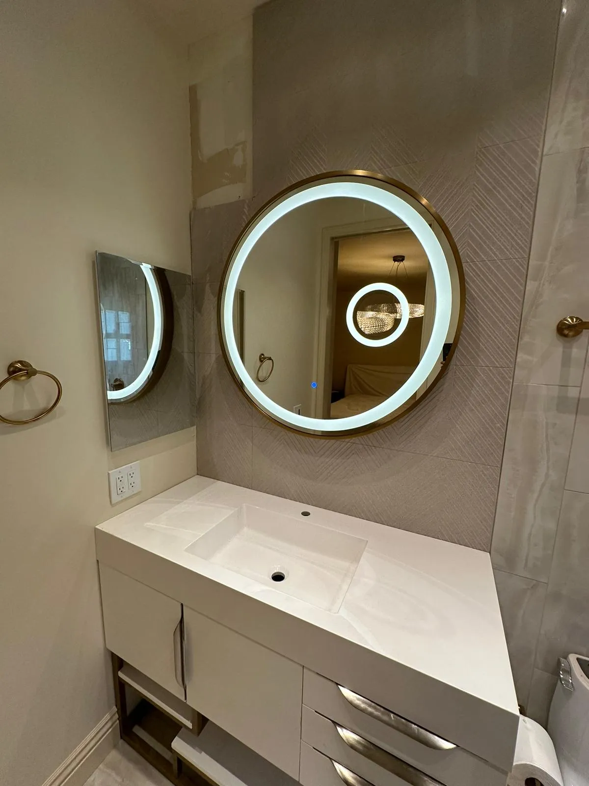 Backlit round mirror with gold frame over white floating vanity. Martinez Electric Corp